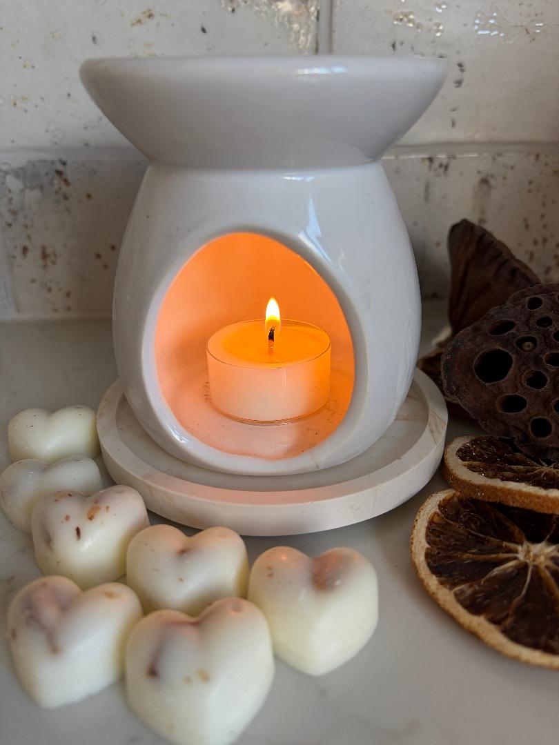White wax melt burner with a lit candle inside, surrounded by heart-shaped wax melts and dried flowers