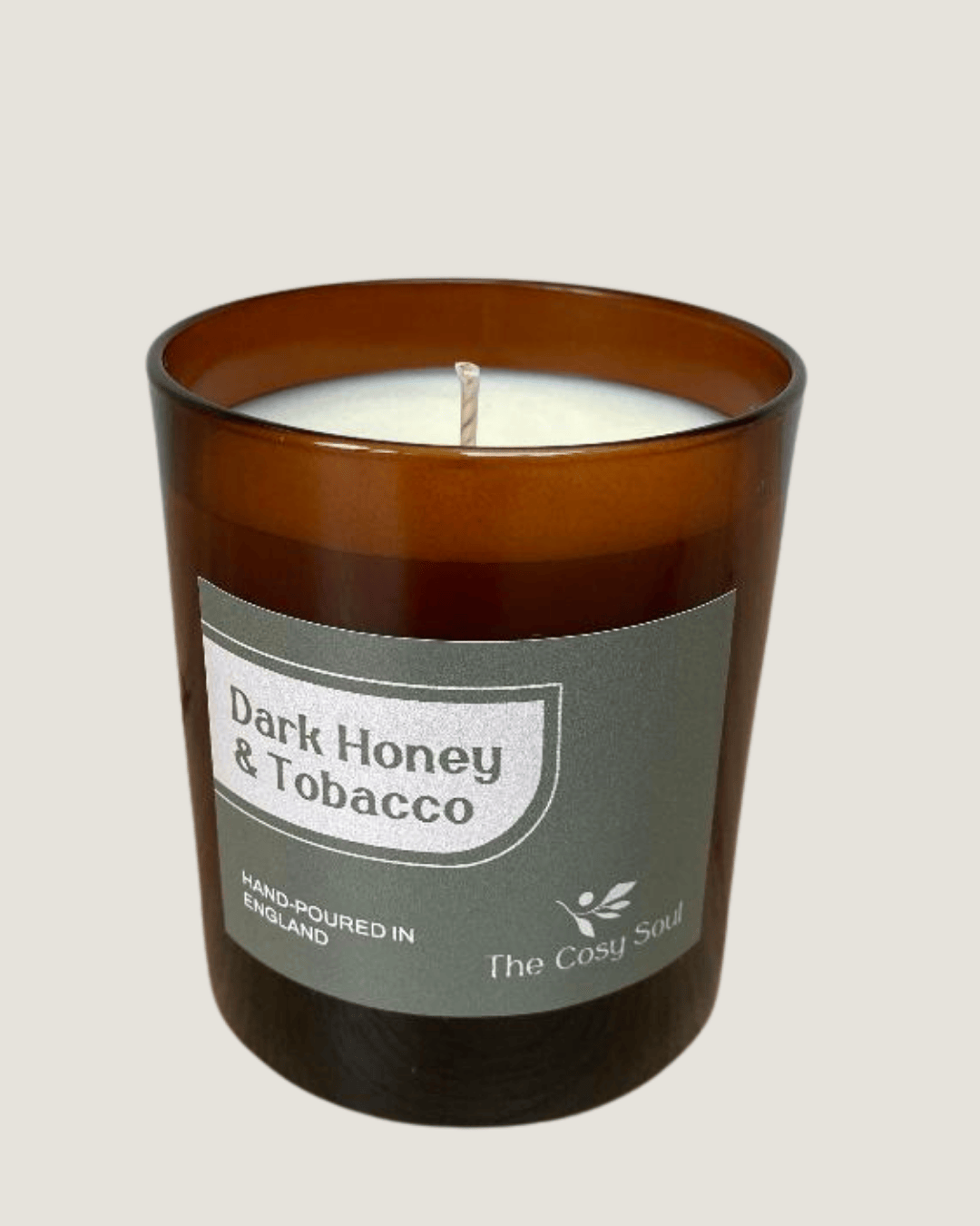 Candle in a brown glass jar labelled 'Dark Honey & Tobacco' manufactured by 'The Cosy Soul' on a beige background