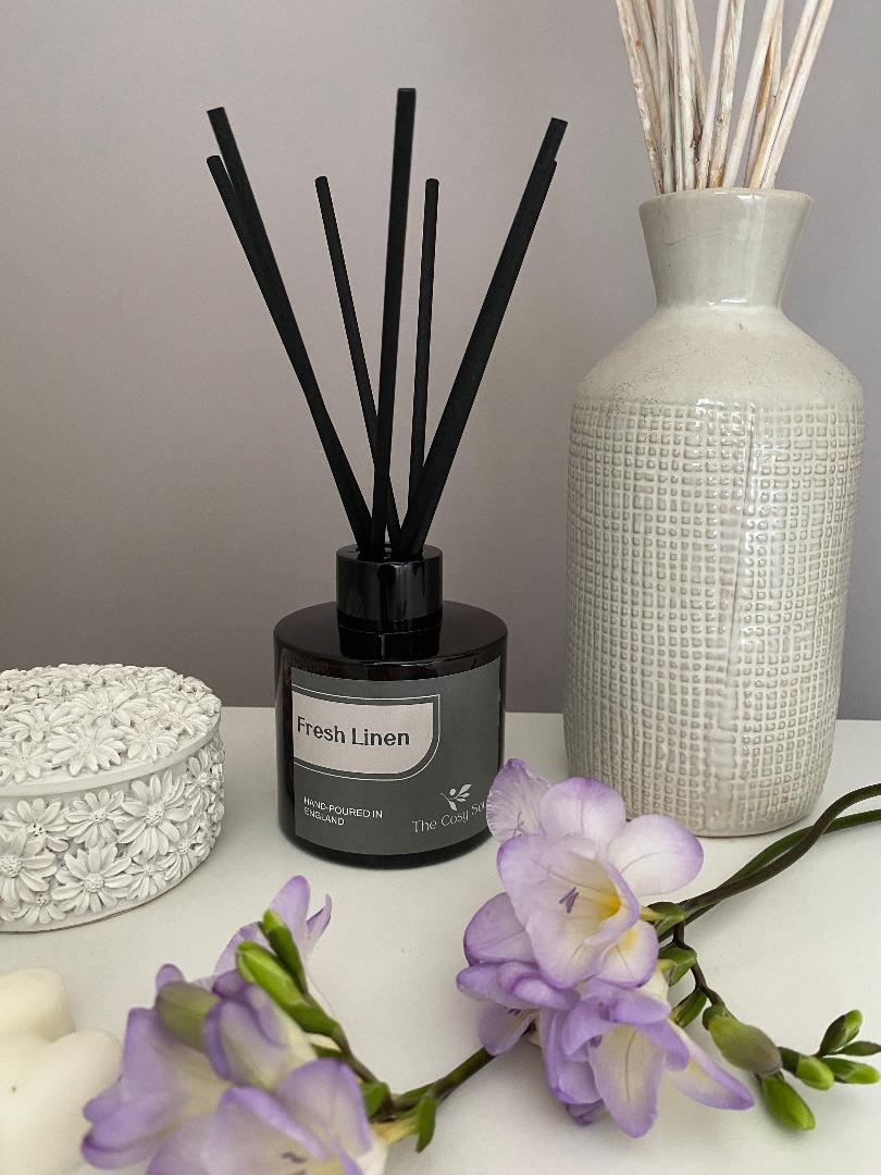 Fresh Linen Diffuser with black reeds, white textured vase, and purple freesia on a light surface.