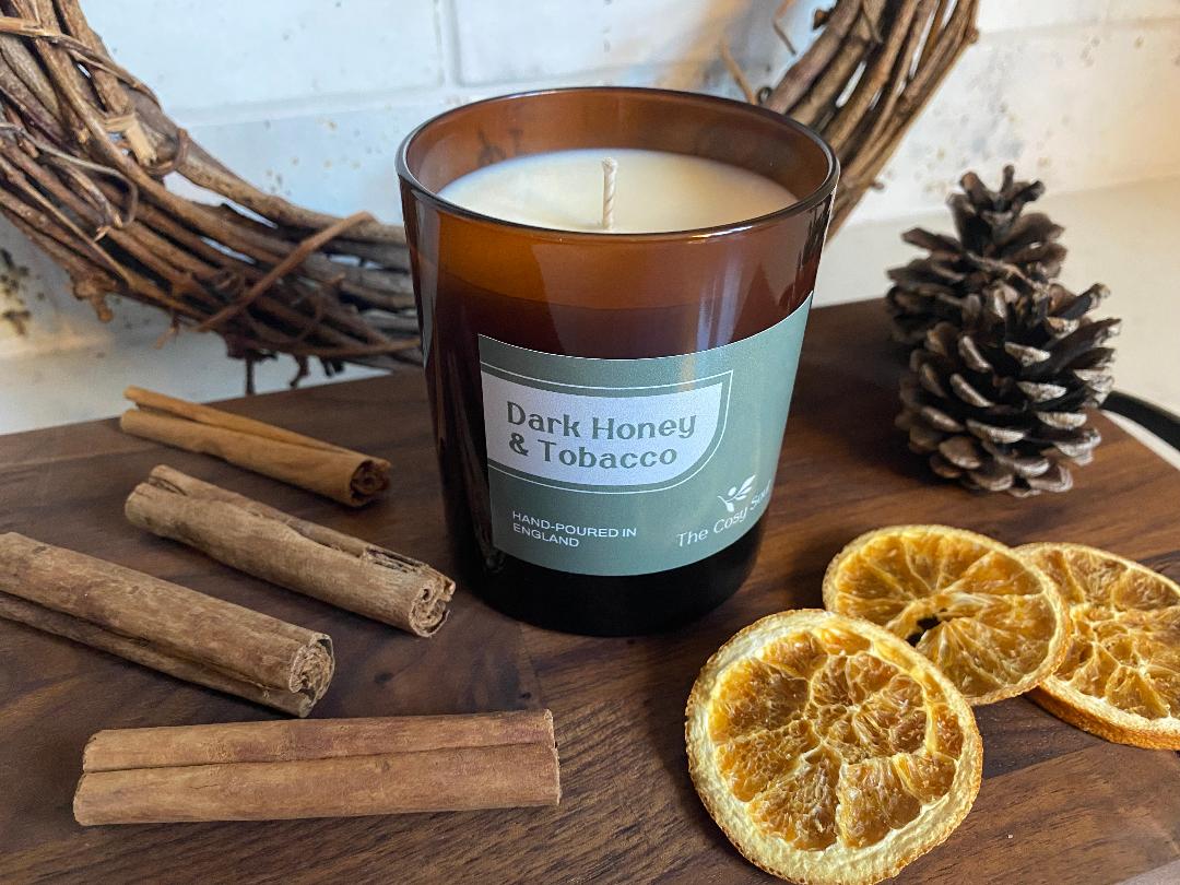 The Cosy Soul Candle labelled 'Dark Honey & Tobacco' surrounded by cinnamon sticks and dried oranges on a wooden surface.