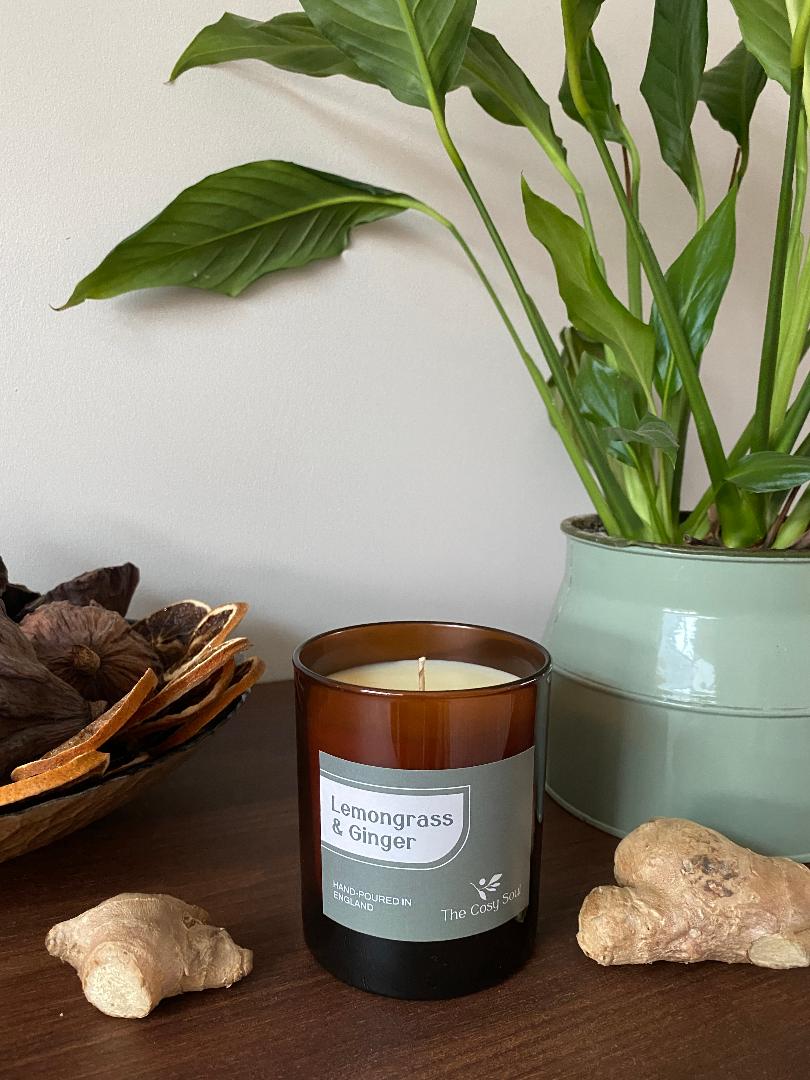 The Cosy Soul Candle fragranced Lemongrass & Ginger on a wooden surface with a plant and ginger root in the background.