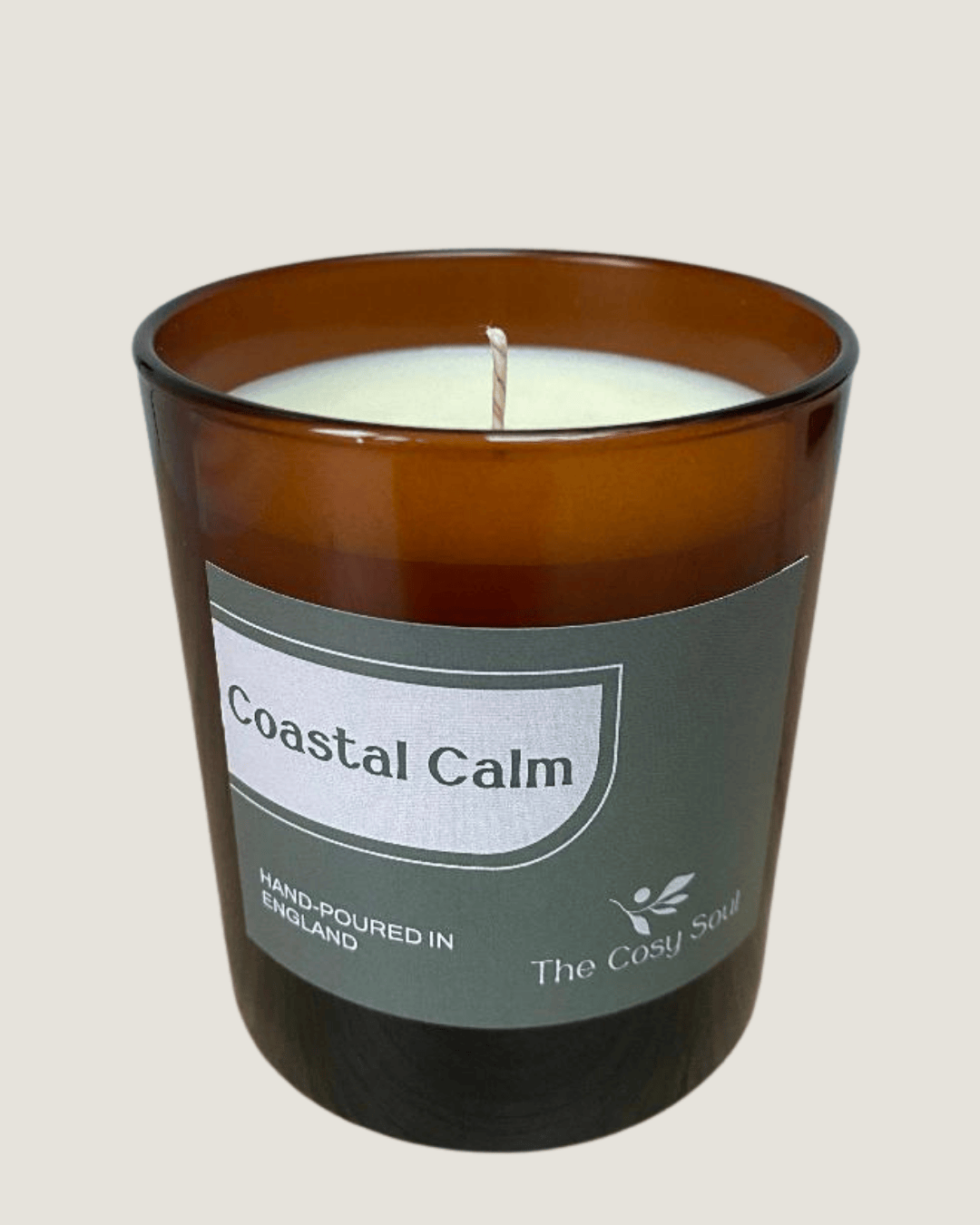 Candle in an amber glass jar labeled 'Coastal Calm' by 'The Cosy Soul' on a light beige background.