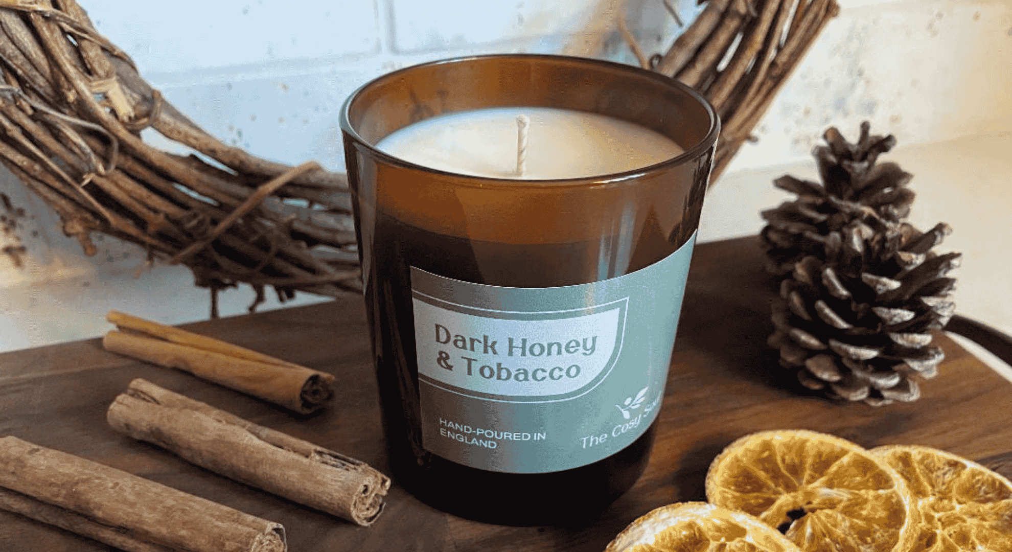 Candle labelled Dark Honey & Tobacco on a wooden surface with cinnamon sticks and pine cones.