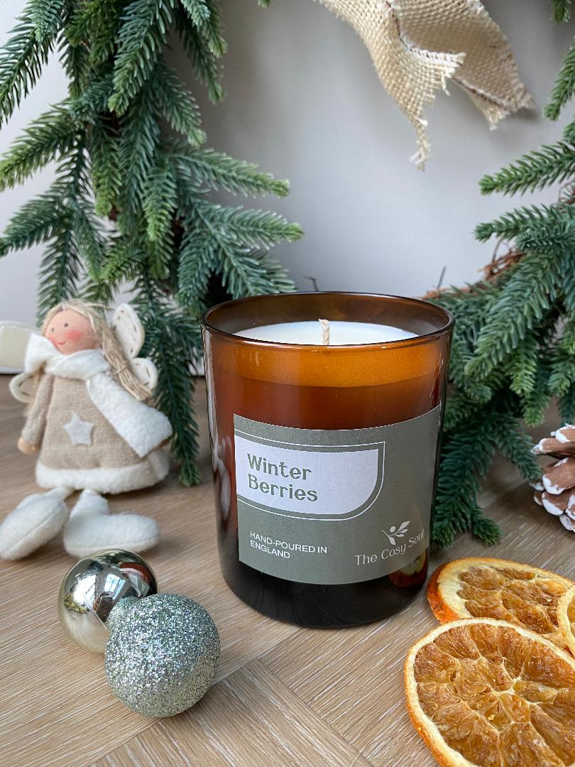 The Cosy Soul Candle labelled 'Winter Berries' with Christmas decor including an angel figurine, ornaments, and dried oranges.
