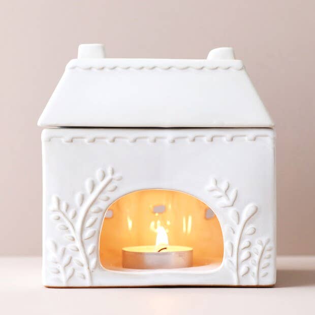 White ceramic house-shaped wax-melt burner with a lit candle inside on a beige background